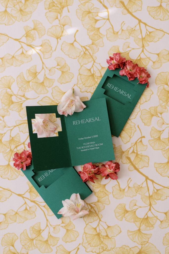 Colorful and unique same-sex wedding rehearsal dinner invitation flatlay on gold gingko linen with colorful florals. The invitation is solid green with a cutout with the dinner details in a modern serif font. The gold linen background complements the design, while the colorful florals add a pop of color and texture. Perfect for a fun and modern same-sex wedding celebration