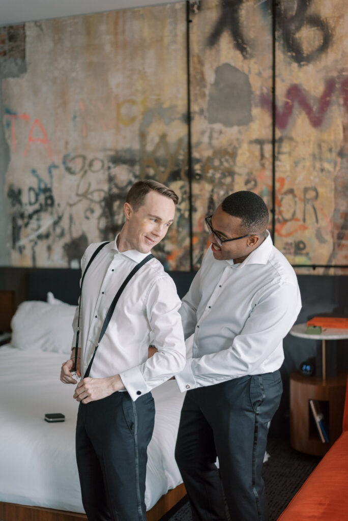 Two grooms-to-be on their wedding day preparing together in an elegant art deco suite at Hotel Haya in Ybor City, Tampa. The couple is seen standing in front of a graffiti wall helping each other with their suspenders. The room has the original wall graffiti and is decorated with decorated with navy red and white details, and large windows let in natural light. Photographed by Kelsey Halm Photography