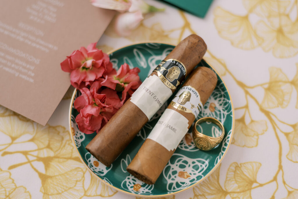 Flatlay of two cigars on a green ceramic floral dish with a gold ring and colorful florals, resting on a luxurious gold gingko linen background