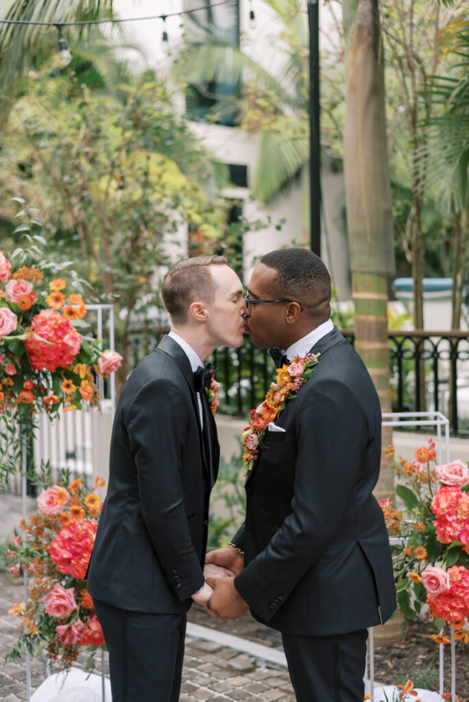 Two grooms, wearing matching custom black tuxes from Indochino, kissing after exchanging their vows. They are an interracial couple, and have vibrant floral pieces, one on his lapel and the other as a pocket garden, adding a pop of color to their ensembles. Palm trees and their ceremony floral pieces create a vibrant background