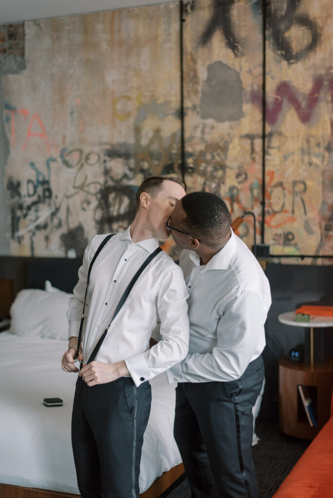 Two grooms-to-be on their wedding day preparing together in an elegant art deco suite at Hotel Haya in Ybor City, Tampa. The couple is seen kissing while standing in front of a graffiti wall helping each other with their suspenders. The room has the original wall graffiti and is decorated with decorated with navy red and white details, and large windows let in natural light. Photographed by Kelsey Halm Photography