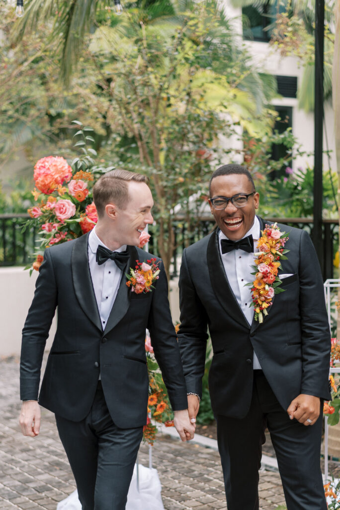 Two grooms, wearing matching custom black tuxes from Indochino, walk hand in hand away from their wedding ceremony. The grooms have vibrant floral pieces, one on his lapel and the other as a pocket garden, adding a pop of color to their ensembles. They are both laughing