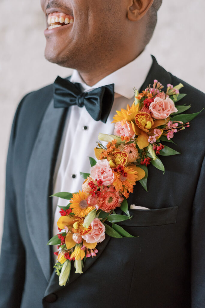 A close-up photograph of a groom's lapel featuring a vibrant and colorful floral arrangement of oranges, yellows, and pinks with greenery accents. The groom is laughing and wearing a custom black tuxedo, black bowtie, and white shirt. This image captures the joy and elegance of a wedding day. Perfect for floral and wedding inspiration searches. Photographed by Kelsey Halm Photography