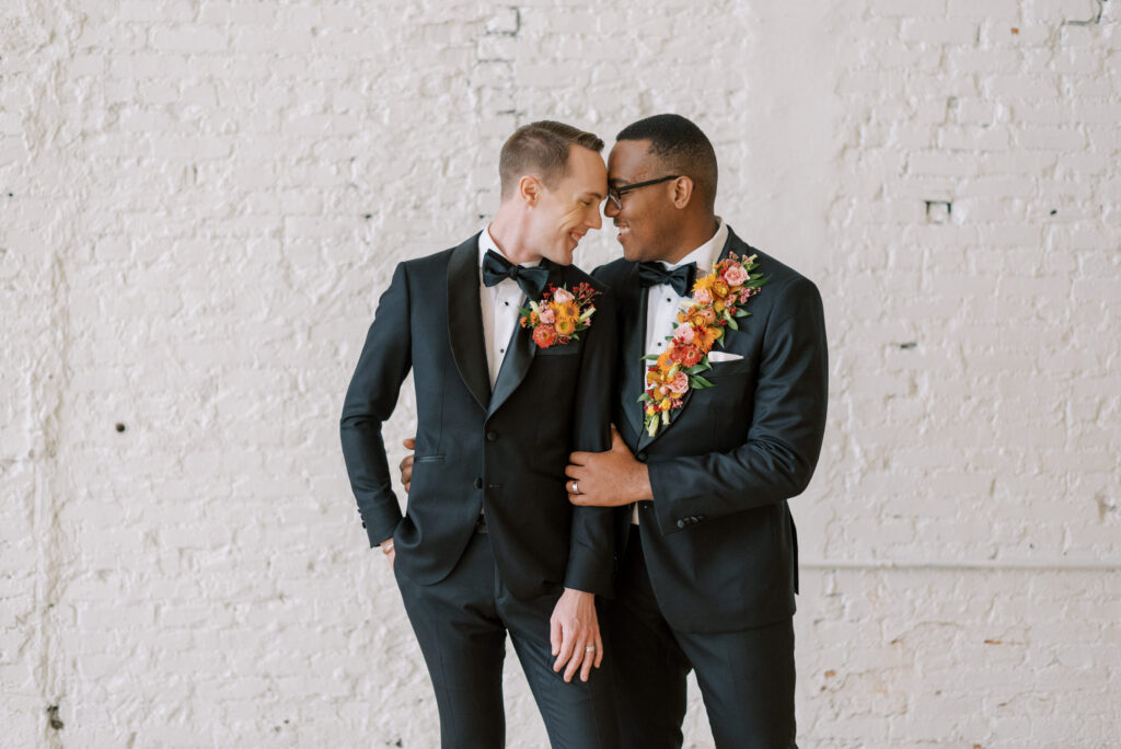 Elegant same-sex wedding photo of two grooms standing close together with unique black tuxedos adorned with vibrant floral lapels and pocket squares at Hotel Haya, beautifully captured by Kelsey Halm Photography