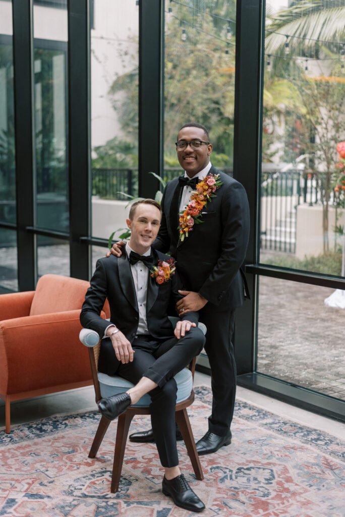 Two grooms are captured in an elegant portrait on their wedding day in the modern eclectic lounge at Hotel Haya. One groom sits in a chair while the other stands beside him, holding his shoulder. Both grooms are dressed in custom black tuxes with black bowties and adorned with vibrant floral pieces, one being a full lapel piece and the other a pocket garden. The florals consist of shades of orange, pink, and green, adding a pop of color to the monochromatic attire.