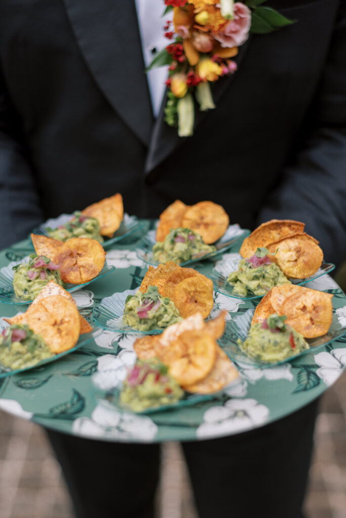 Close-up photo of a groom holding a custom platter of appetizers during cocktail hour at a wedding reception. The platter matches the green and white table napkins of the reception decor. The appetizers are guacamole and plantain chips, and they look delicious and colorful.