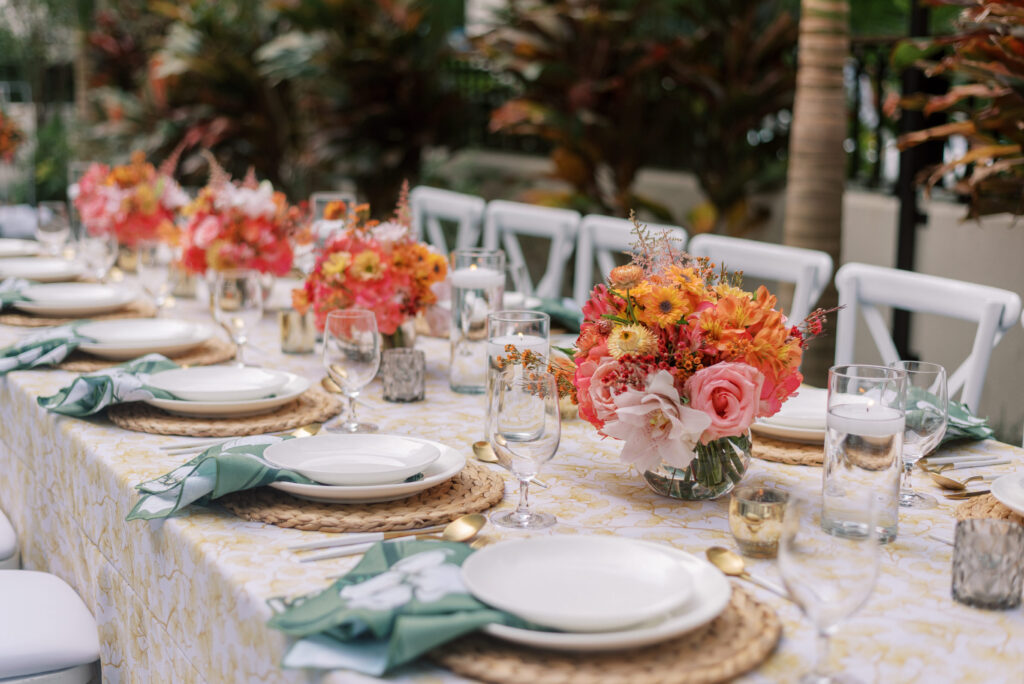 A stunning outdoor wedding reception table setup in the courtyard of Hotel Haya in Ybor City. The long table is adorned with vibrant and voluminous floral centerpieces in shades of orange, pink, and yellow. The table linen is a beautiful gold gingko pattern, and the napkins feature green with white floral prints. The table setting includes rattan chargers, elegant white plates, and unique peacock glasses, creating a perfect blend of modern elegance and fun