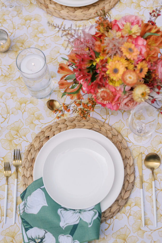 A close-up photograph of a single place setting on a long wedding reception table, set up in the outdoor courtyard of the Hotel Haya in Ybor City. The plate is white and rests on top of a rattan charger, with vibrant floral pieces in orange, pink, and yellow creating a stunning centerpiece. The napkin is green with white floral designs, placed on top of a gold gingko-patterned tablecloth. Gold flatware with white handles complete the elegant place setting