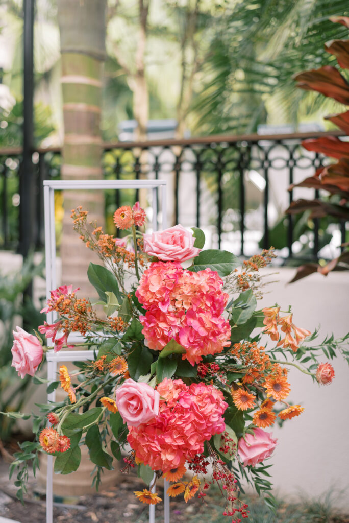 Vibrant floral installations on white frames for wedding ceremony. The blooms feature bold and bright hues of orange, pink, and greenery creating a voluminous display.
