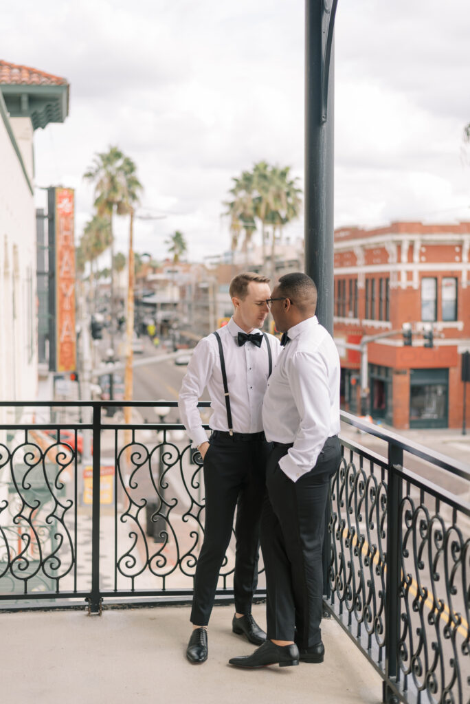 Two grooms are captured in a black and white photo standing on the historic iron balcony at Hotel Haya in Ybor City, Tampa. They stand forehead to forehead, sharing a quiet moment before their wedding ceremony. Both grooms are dressed in white shirts, black bowties, and black suspenders paired with black tux pants.