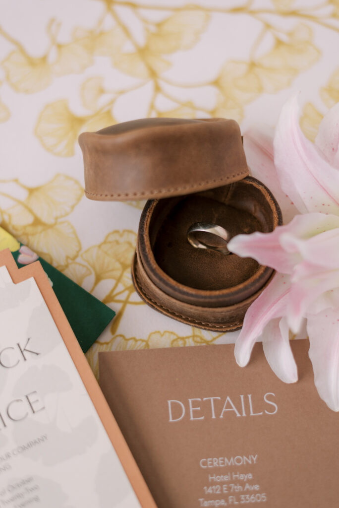 Handcrafted leather ring box displaying groom's ring photographed in a flatlay on a luxurious gold gingko linen with a golden ring and vibrant florals, perfect for a stylish and elegant wedding ceremony.