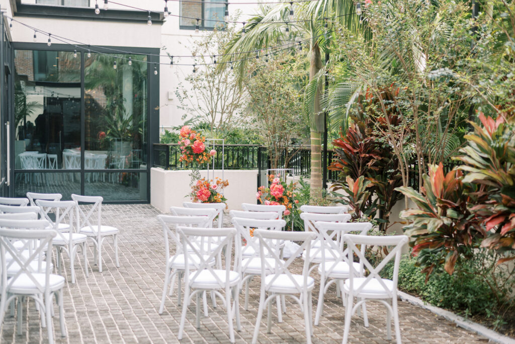 Intimate wedding ceremony setup at Hotel Haya in Ybor City Tampa, Florida. The brick courtyard features white chairs arranged in a curved aisle, leading to a stunning ceremony site with two asymmetrical floral installations. The vibrant and voluminous pink, orange, yellow and green flowers create a colorful and eye-catching display against the brick pavers. 