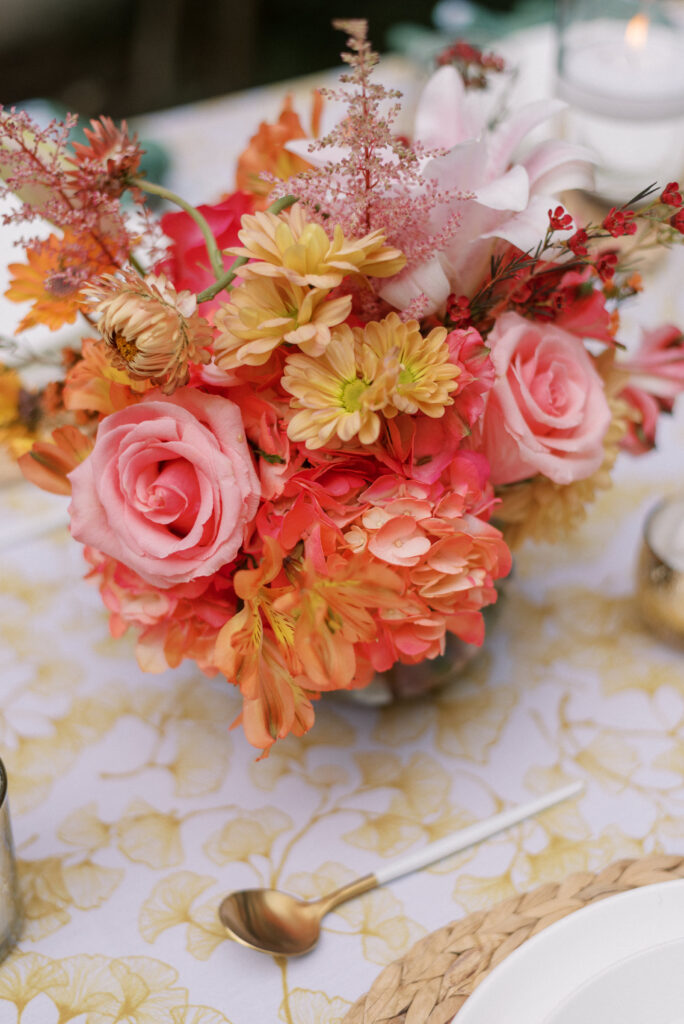 A close-up of a vibrant orange and pink floral centerpiece placed on a reception table covered with a golden gingko patterned linen. The flowers are in full bloom, creating a lively and cheerful atmosphere for the guests.