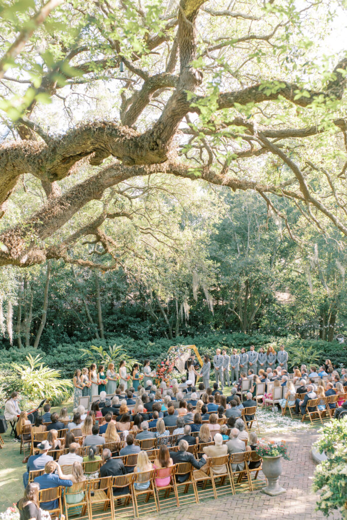 Colorful wedding ceremony under live oaks in the courtyard of the Thomas Bennett House with custom cane backed chairs, brick pavers, and sun shining through leaves. Photographed by Destination Wedding Photographer Kelsey Halm Photography