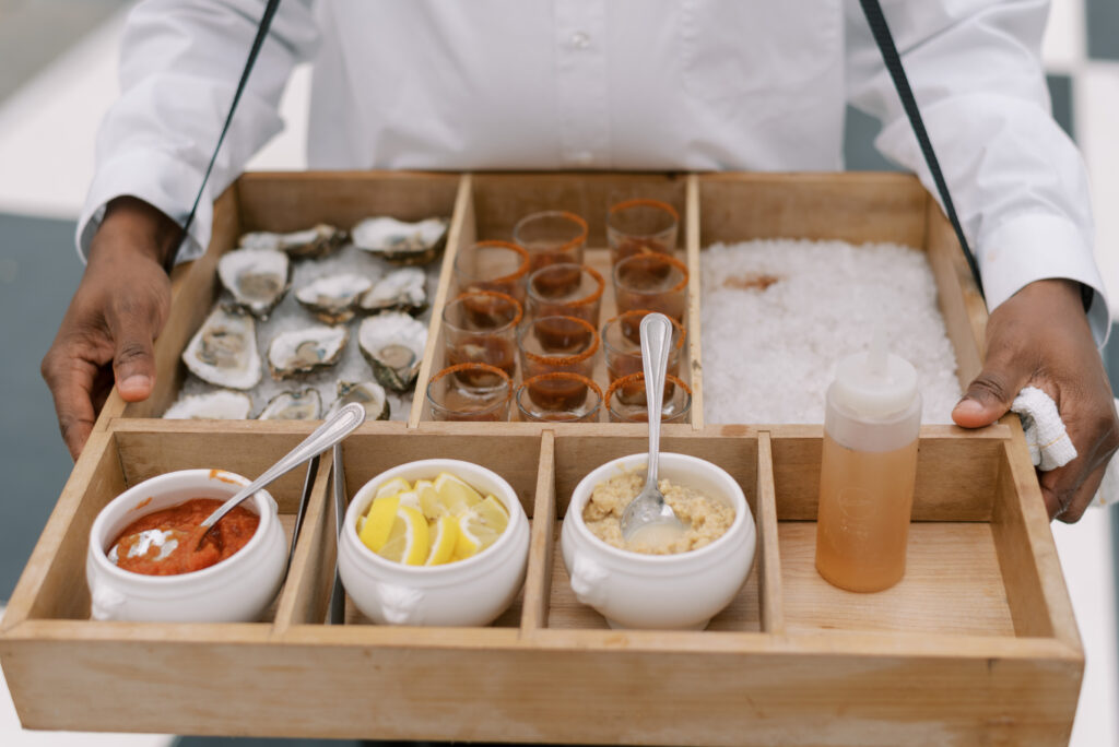 A waiter holding a tray of delicious oyster shooters served as small bites at a beautiful outdoor wedding reception at Lowndes Grove in Charleston, South Carolina. The waiter is wearing a white shirt and the photo is captured by destination wedding photographer Kelsey Halm.