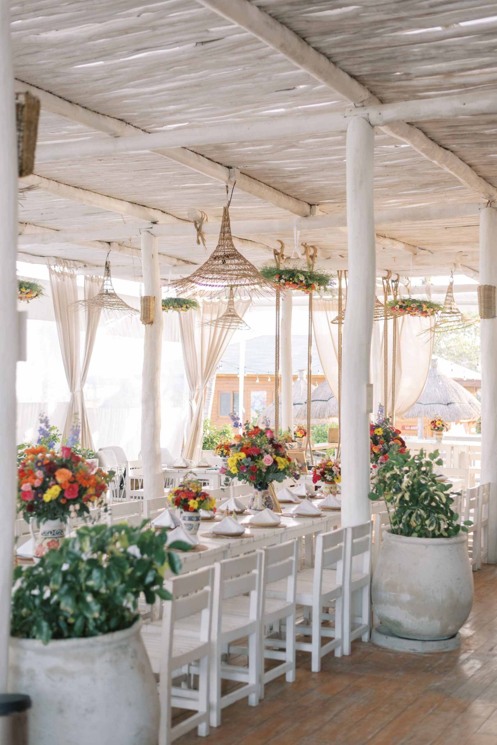 A beachside rehearsal dinner setup at a white and driftwood terrace in Akumal, Mexico. Colorful flowers and traditional Mexican pottery adorn the table as guests gather around to celebrate. The turquoise waters of the Caribbean sea provide a stunning backdrop. This photograph was captured by destination wedding photographer Kelsey Halm.