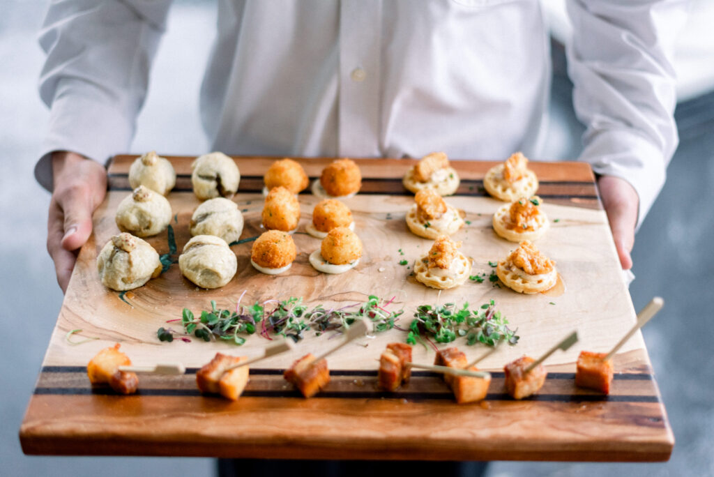 An elegant waiter wearing a crisp white shirt holds a butcher board tray with an assortment of delicious small bites. The appetizers include crabcakes, mini chicken and waffles, pork belly.  Perfect for any occasion, these delectable hors d'oeuvres are sure to impress your guests.