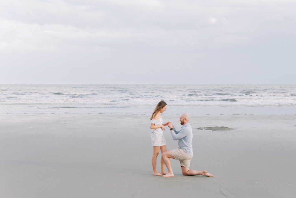 man puts ring on his fiancé's finger while he's down on one knee on the beach