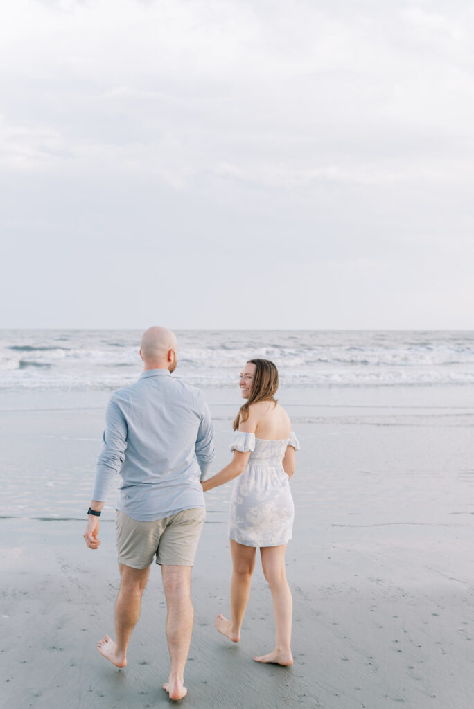 engaged couple holding hands walk towards the ocean 