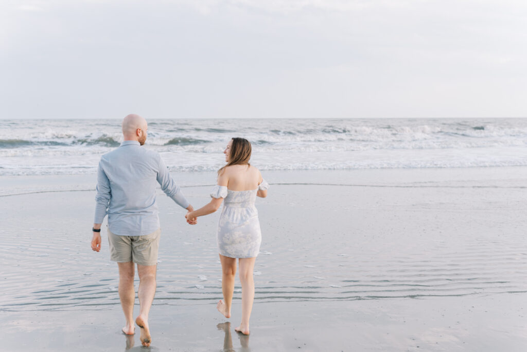 engaged couple holding hands and smiling at each other while they walk into the water at the beach