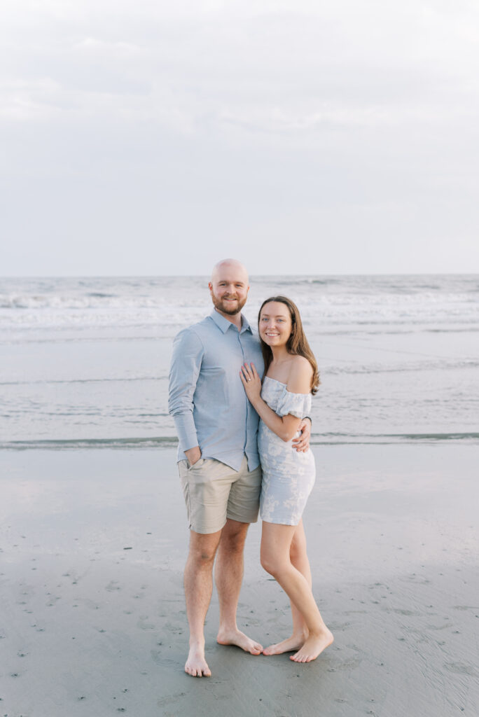 engaged couple wearing blue smiling at the camera in front of the ocean at kiawah island beach 