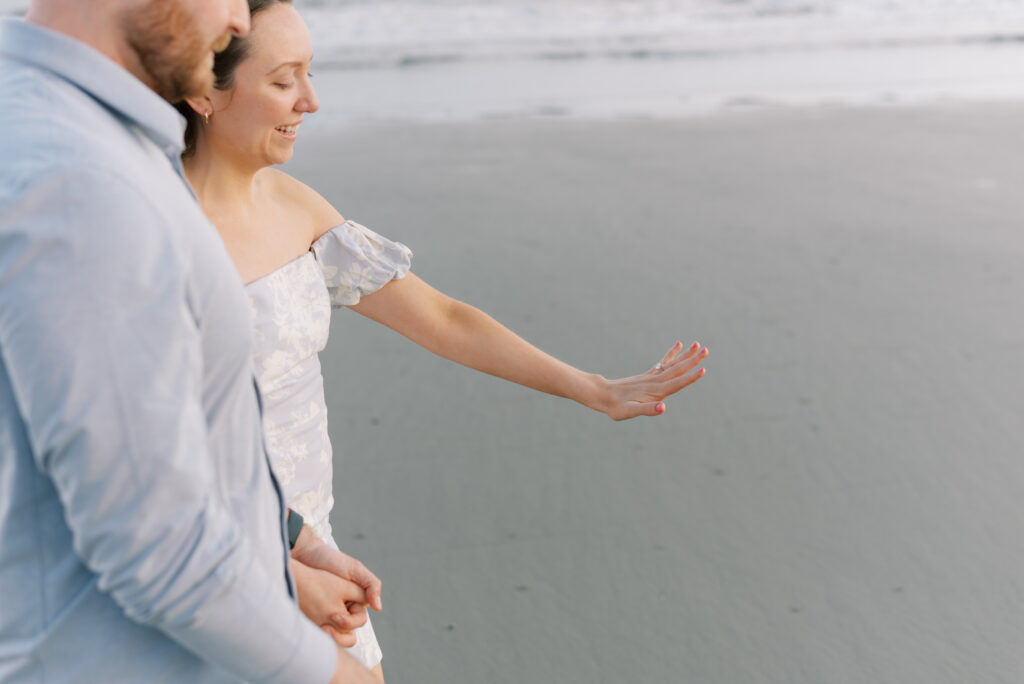 woman looks at her new ring while holding her fiancé's hand on the beach