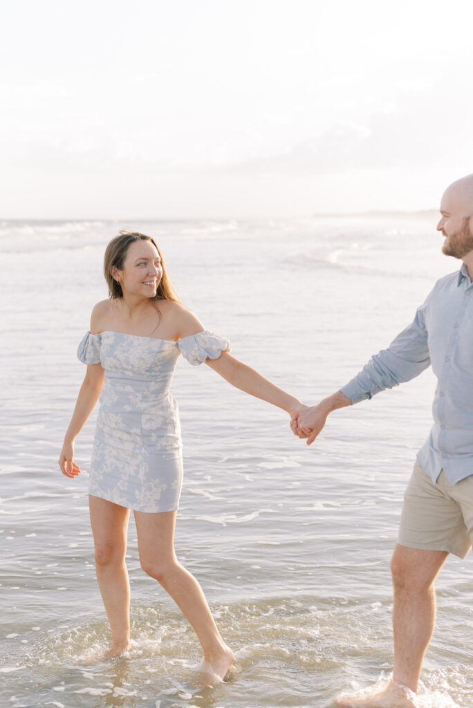 couple holds hands and walks through water at sunset