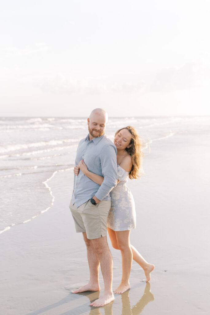 woman hugs her fiance from behind while standing in the shallow waves of the ocean