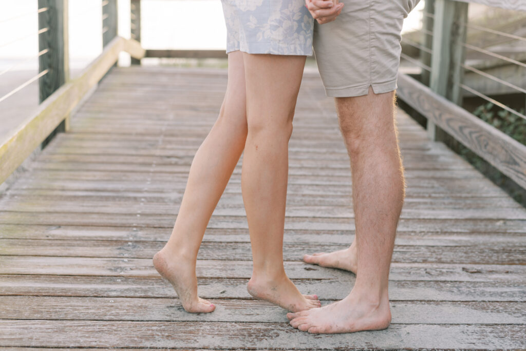 close up of man and woman's legs standing on a beach boardwalk while they hold hands and kiss
