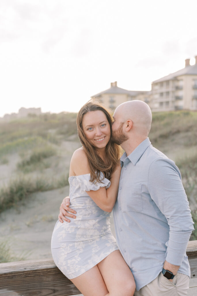 woman sits on a railing on a beach boardwalk while her fiancé hugs her