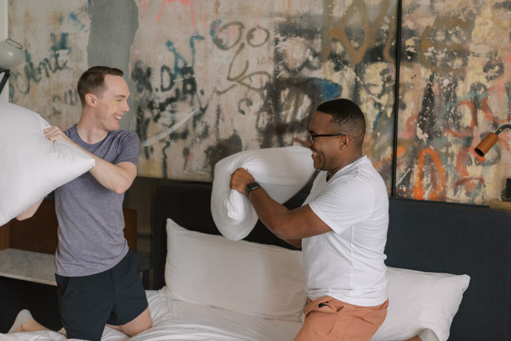 gay husbands have a pillow fight and laugh on their hotel bed in front of graffiti wall