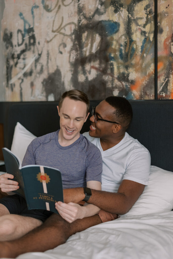 man reads to his husband while sitting together on their hotel bed