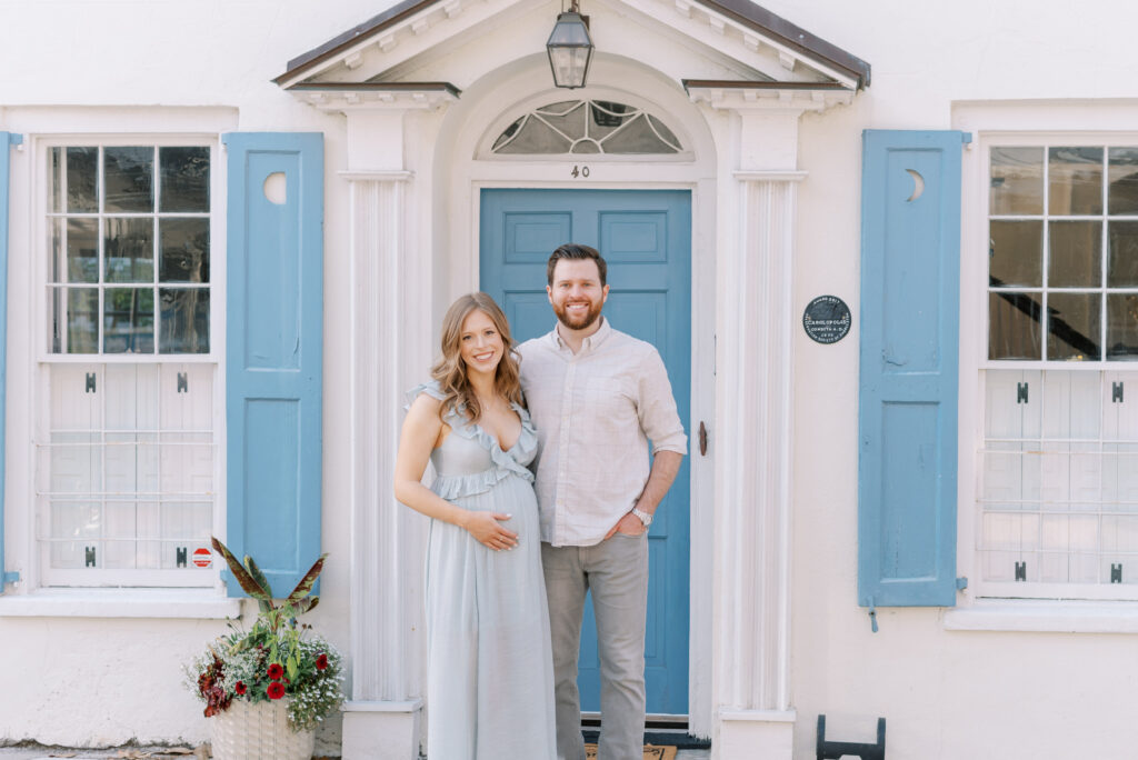 white house with blue shutters with a pregnant couple smiling in front of it