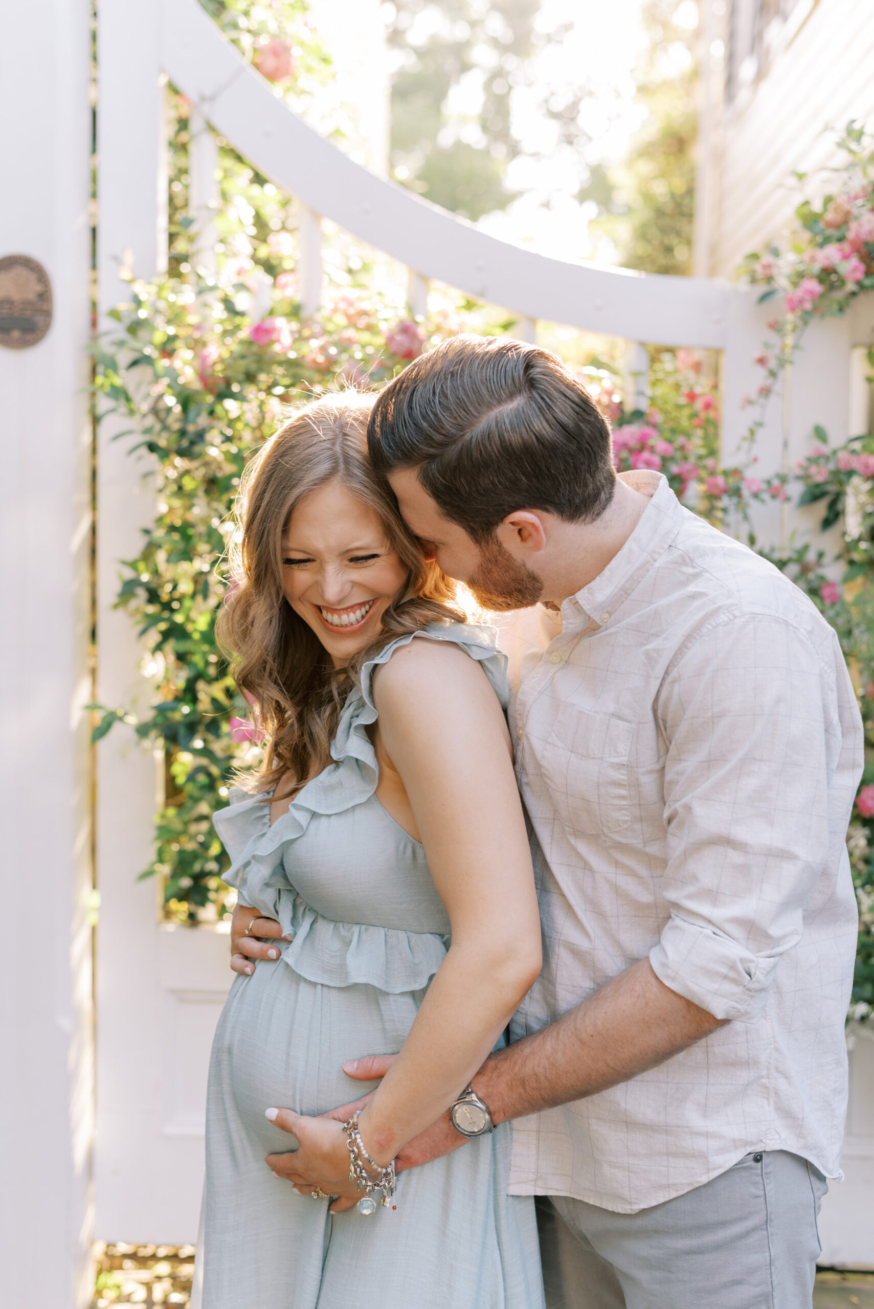Man kisses his wife on the cheek in front of a gate covered in flowers maternity session