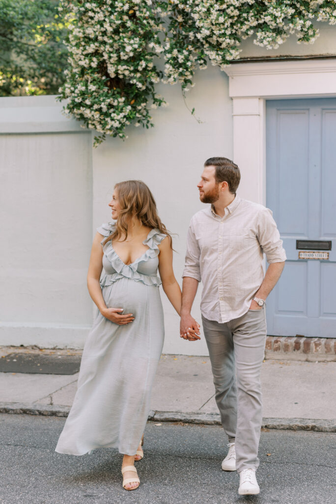 man and woman walk across a street during their maternity photo session in front of a blue door in Charleston