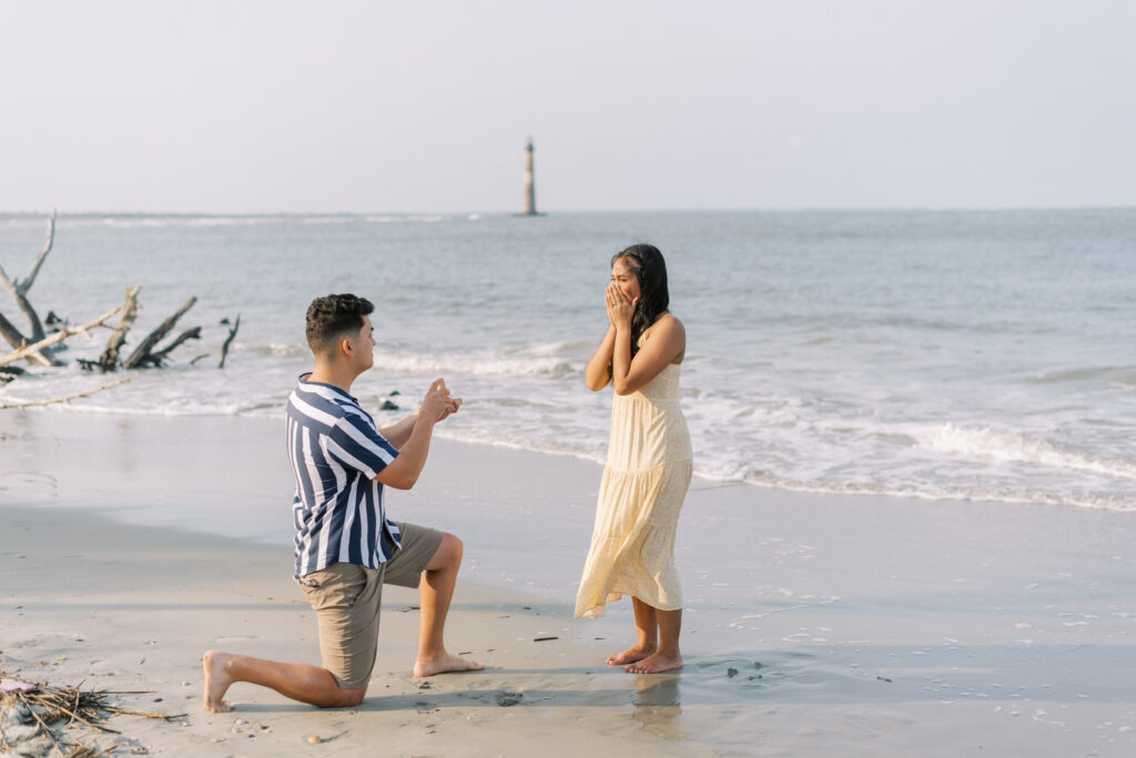 Woman gasps in shock as her boyfriend proposes on Folly Beach with Morris Island Lighthouse in the background