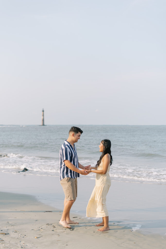 couple hold hands and smile at each other right after a proposal on the beach