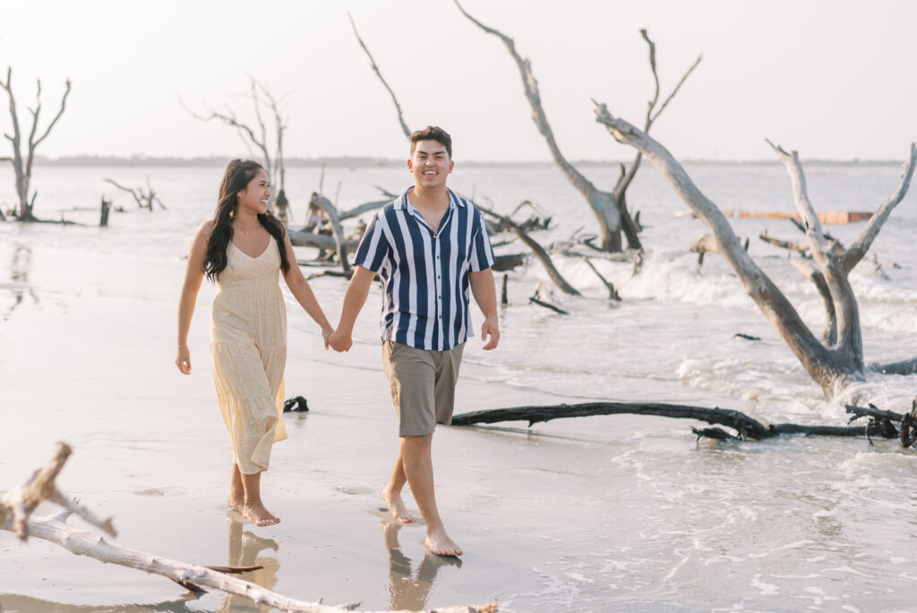 Couple hold hands and walk toward the camera through driftwood on the beach