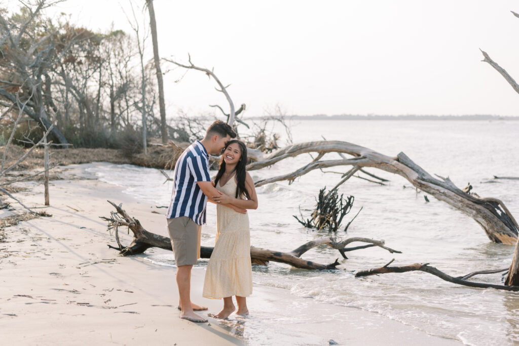 Couple laugh together as man whispers in his fiancé's ear amid driftwood on folly beach
