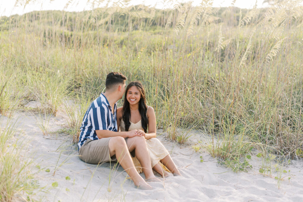Newly engaged couple sit in the sand amid beach grass