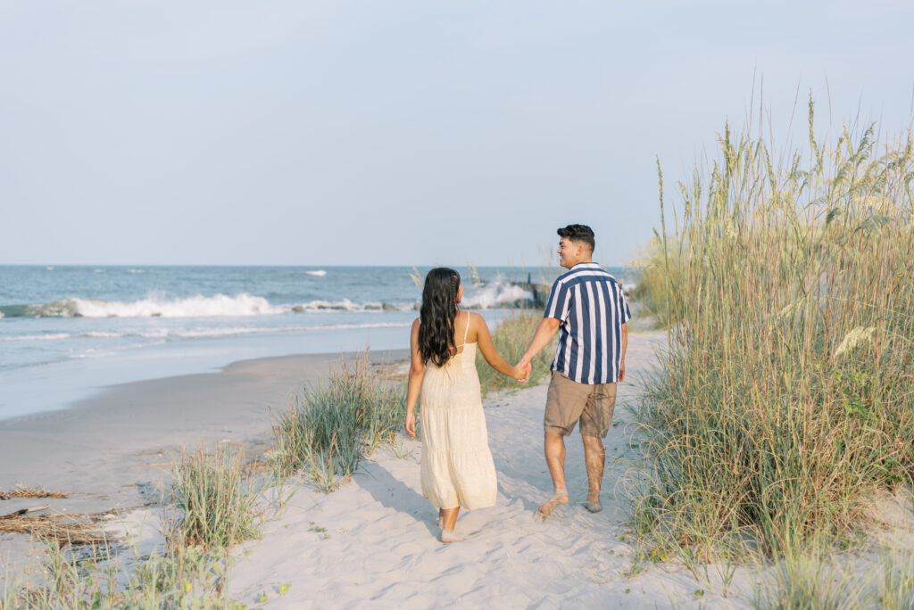 Couple holding hands and walking along a beach path with water in the background