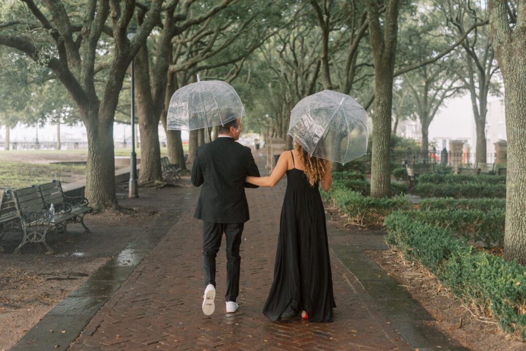 Couple walk together under umbrellas through the Live Oaks in a Charleston park