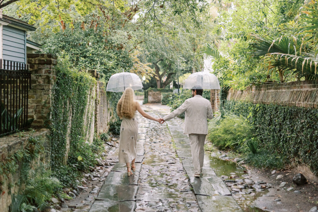 Engaged couple walking down an ivy covered alley in Historic Charleston with umbrellas