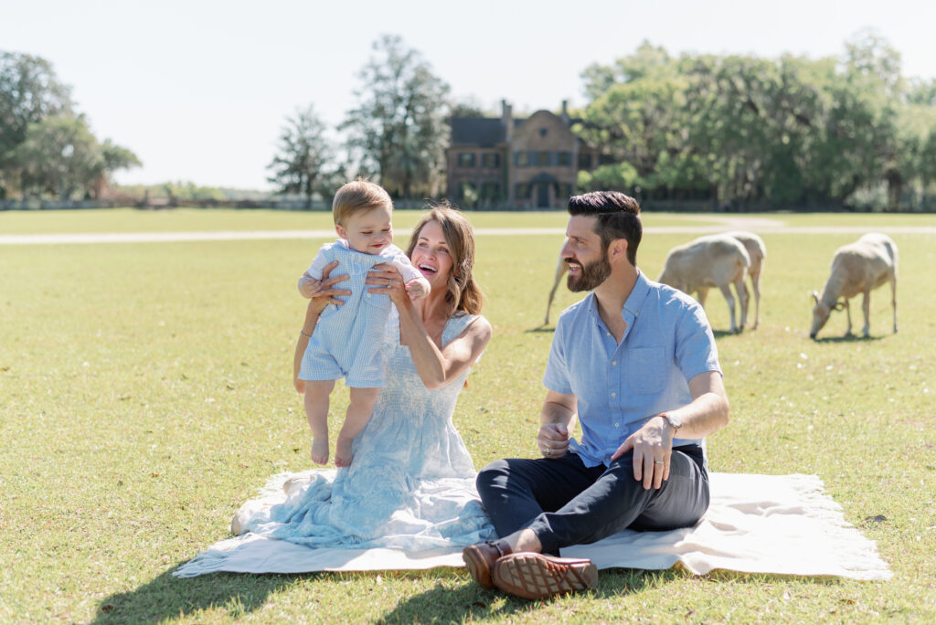 Mother and father laugh with their baby sitting on a blanket at Middleton Place amid sheep