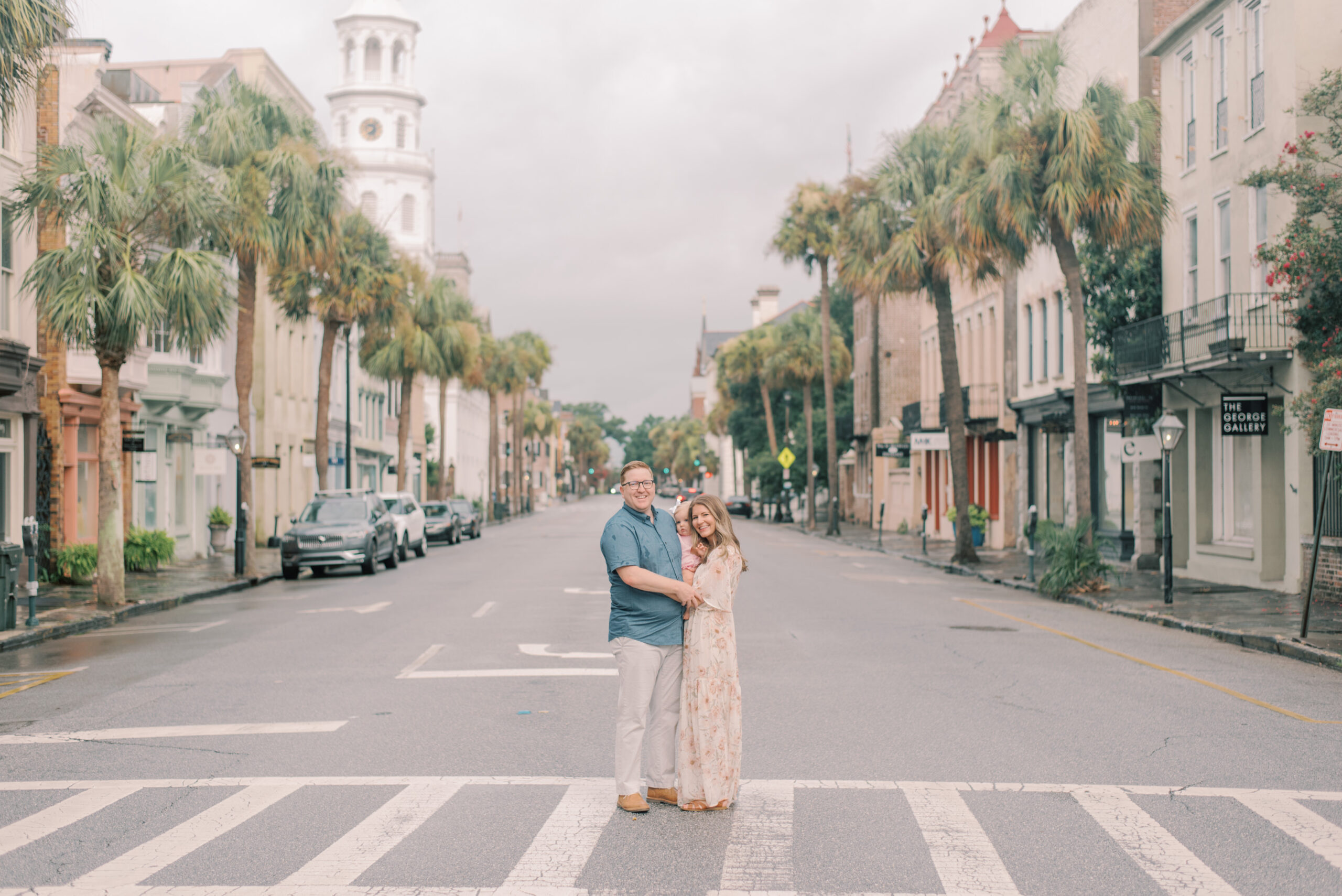 Family of three standing in the middle of Broad St. in Charleston with palm trees and colorful buildings